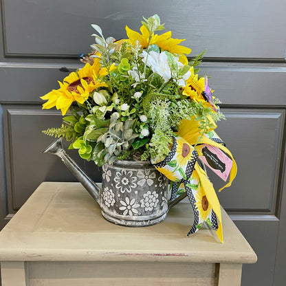 Floral arrangement in a watering can with a decorative ribbon on a wooden surface.