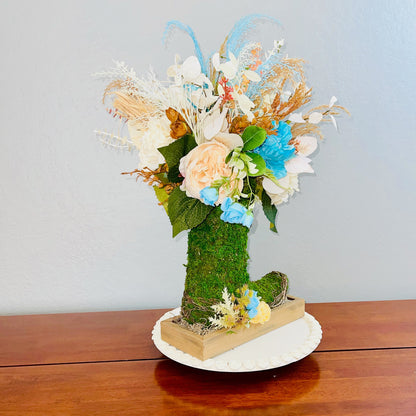 Decorative floral arrangement in a moss-covered boot on a wooden surface with a light blue background