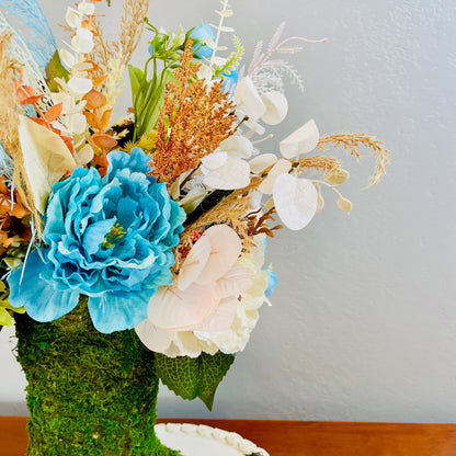 Floral arrangement with blue and white flowers in a moss-covered vase on a wooden surface.