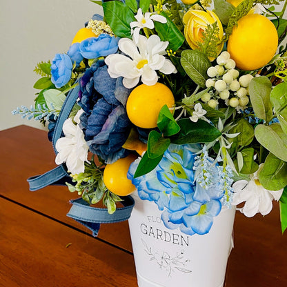 Bouquet of flowers with lemons and blue ribbons in a white vase labeled 'Floral Garden' on a wooden surface.