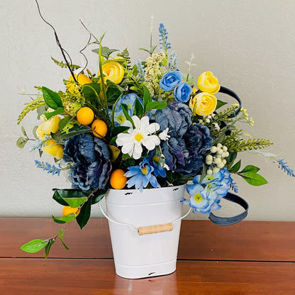 Floral arrangement with blue, yellow, and white flowers in a white bucket on a wooden surface.