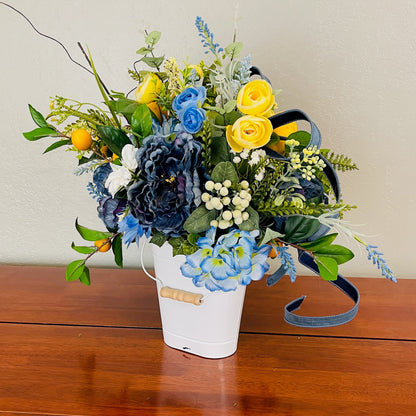 Floral arrangement with blue, yellow, and green flowers in a white bucket on a wooden surface.