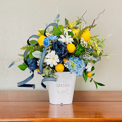Floral arrangement with blue, yellow, and white flowers in a white bucket on a wooden surface.