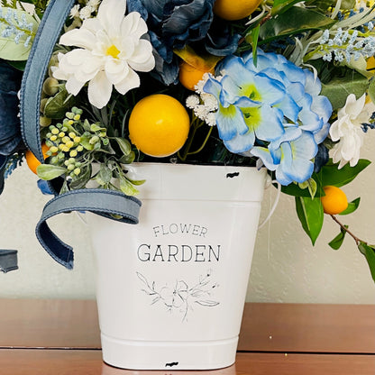 Floral arrangement in a white bucket labeled 'Flower Garden' on a wooden surface.