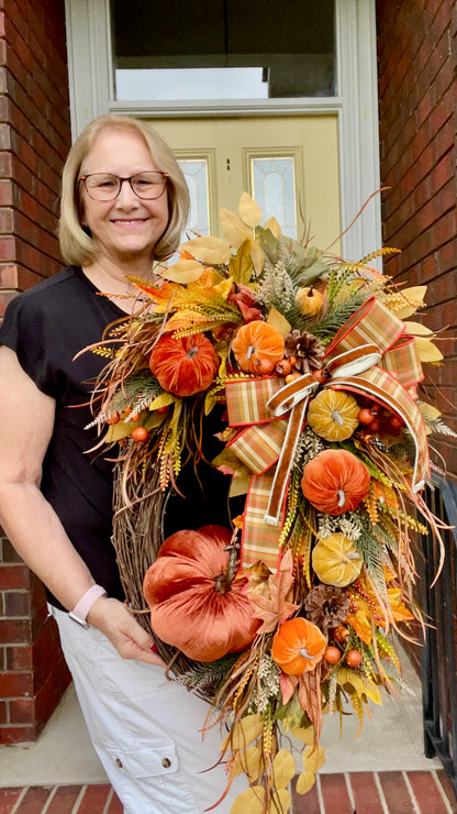 Woman holding a large autumn wreath with pumpkins and leaves in front of a door.
