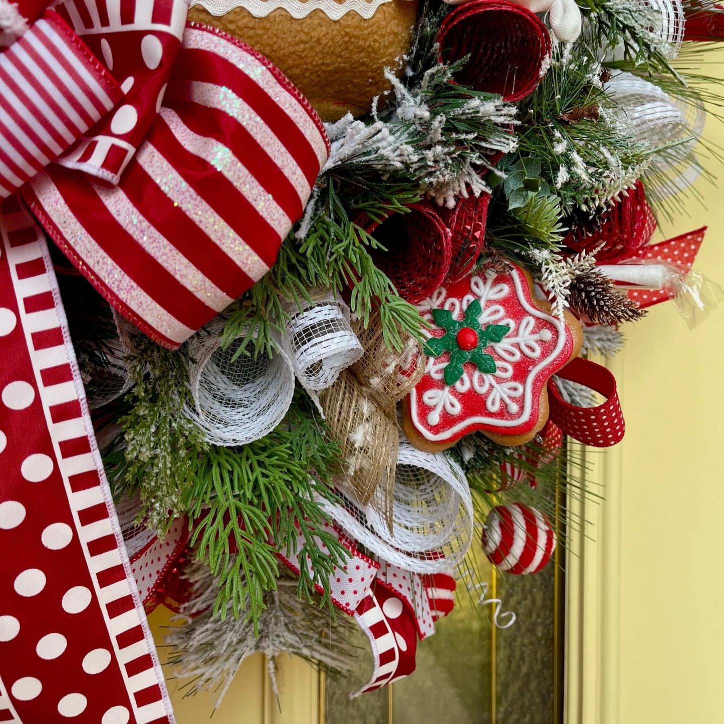 Decorative Christmas wreath with red and white ribbons, greenery, and a gingerbread cookie design on a yellow door.