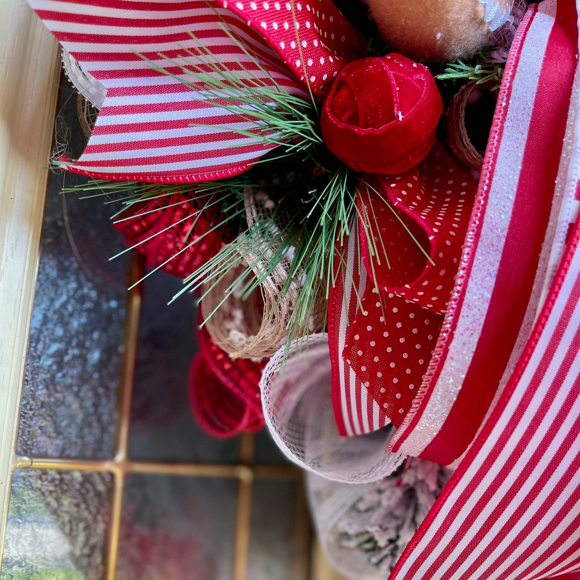 Decorative wreath with red and white ribbons, pine cones, and greenery on a glass door.